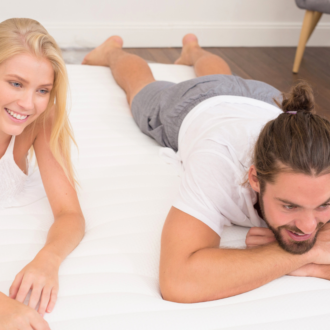 A man and a woman lying on a white mattress topper, with the topper's brand name 'Naptime' visible.