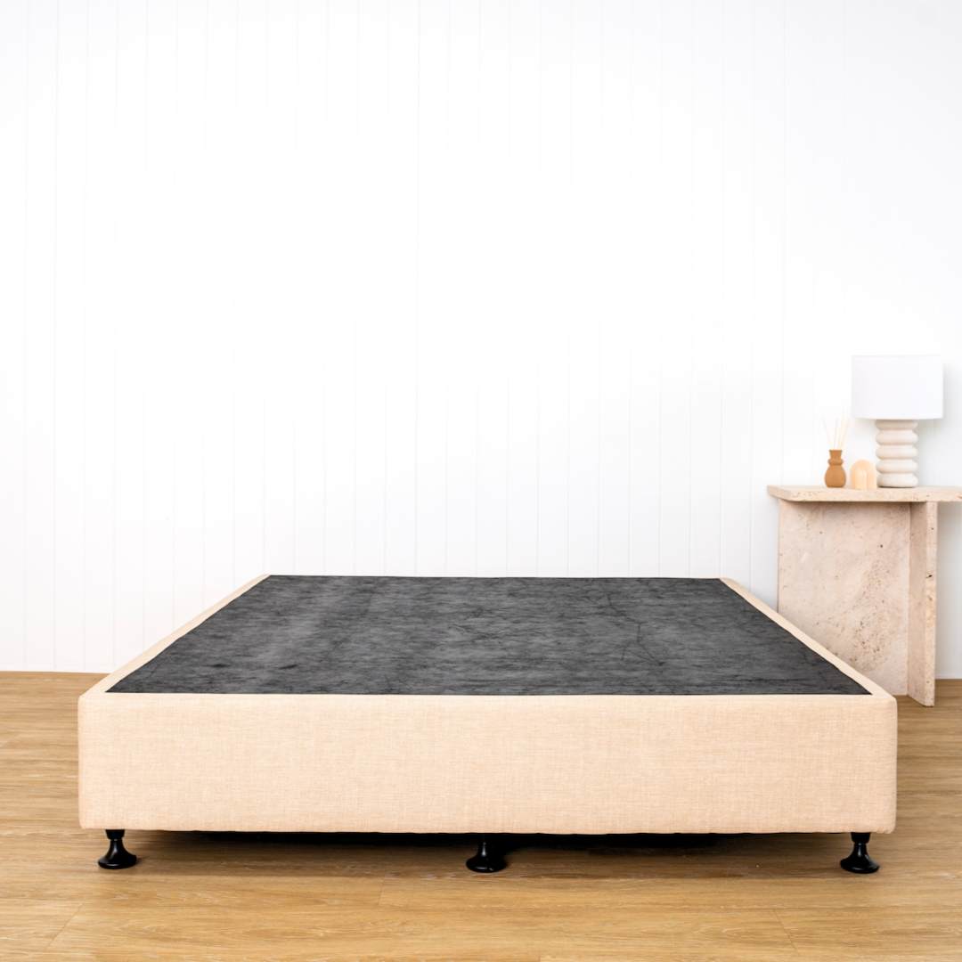 A beige bed base with a textured surface on top, placed on a wooden floor against a white wall, with a small lamp and table to the right.