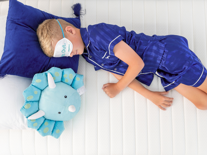 Child in blue pajamas with dinosaur pillow and eye mask on a Naptime bed.