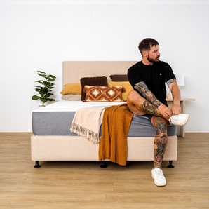 A person sitting on a bed with a medium-firm mattress, which is dressed in a gray mattress protector. The room has a light color scheme with decorative pillows and a blanket.