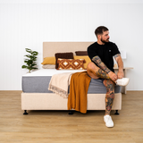 A person sitting on a bed with a medium-firm mattress, which is dressed in a gray mattress protector. The room has a light color scheme with decorative pillows and a blanket.