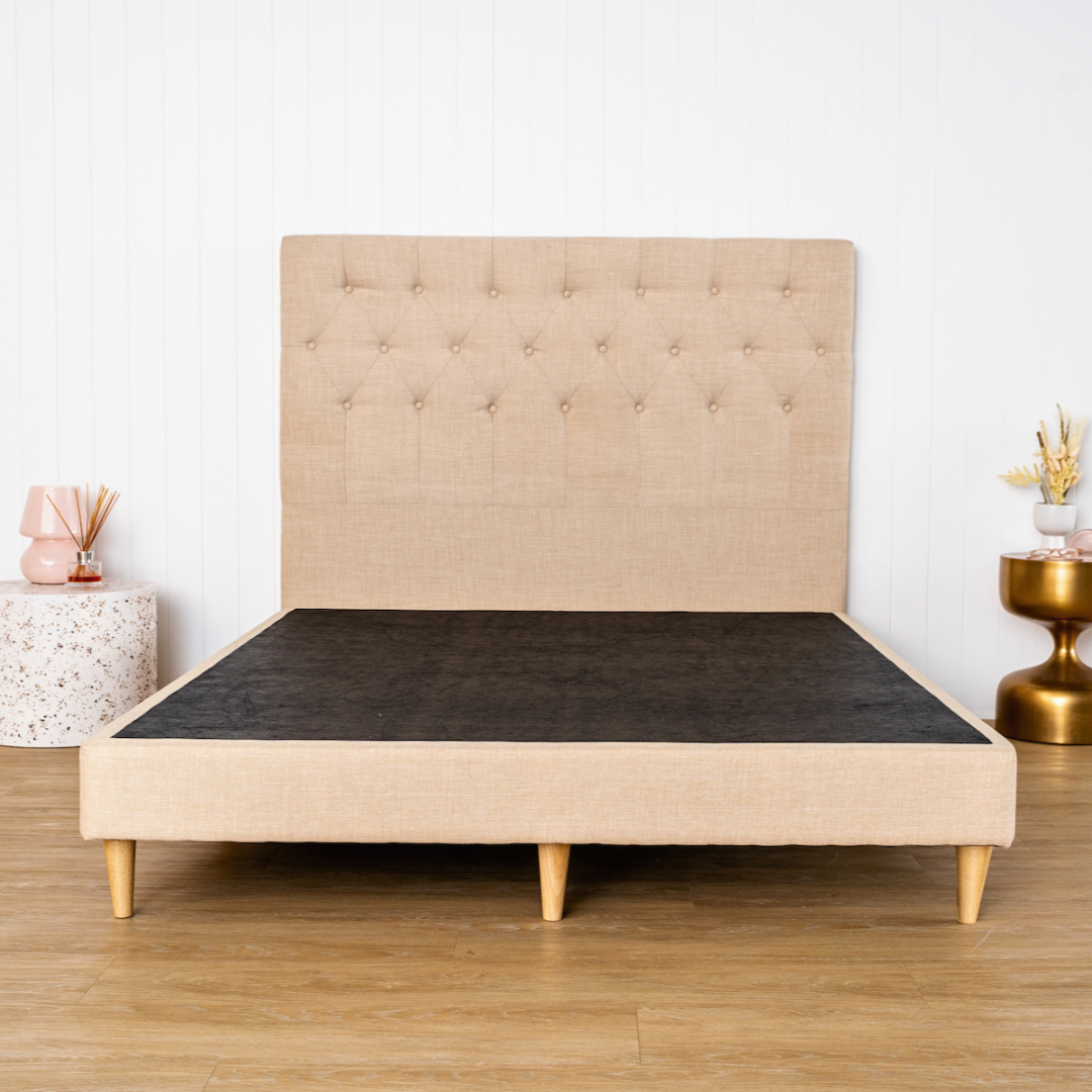 A natural upholstered bed frame with a high headboard and wooden legs on a wooden floor, with decorative objects beside it.l 
