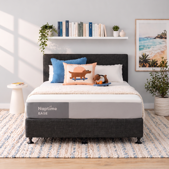 A twin-size mattress in a dark gray upholstered bed frame, styled in a bright, modern bedroom. The mattress features a gray and white panel with the text 'Naptime EASE' printed on it.